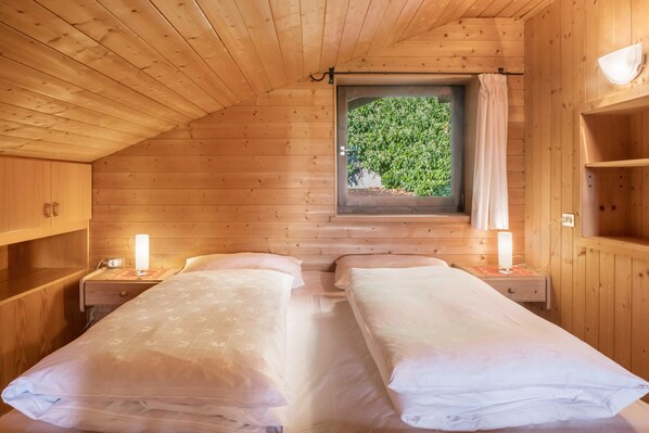 Ein Zimmer in einer Holzhütte mit zwei Betten, einem Fenster mit Vorhang und einem Regal.