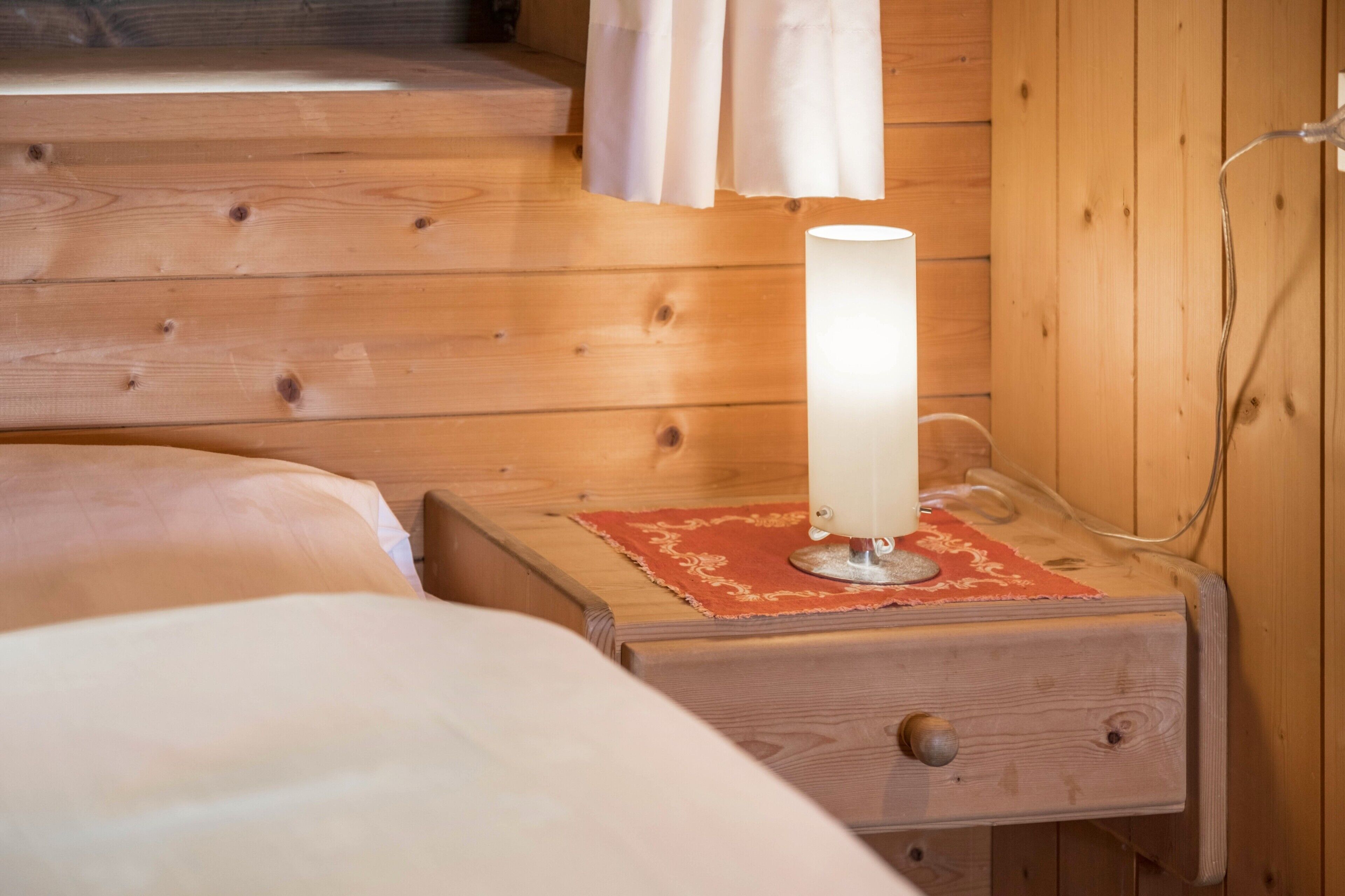 A wooden cabin interior with a bed, a nightstand with a lamp, and a wooden wall.