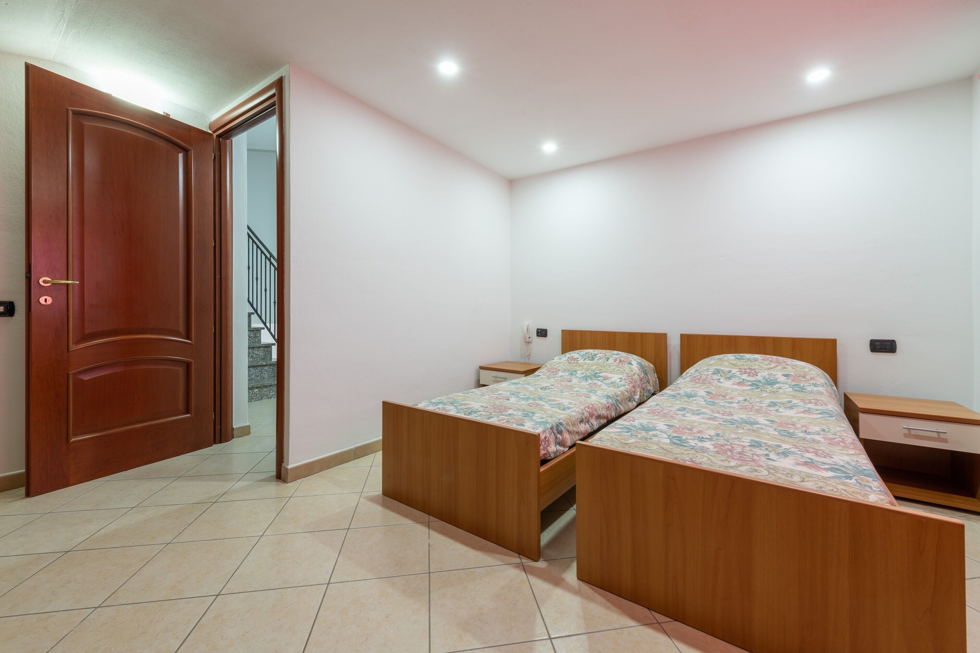 Een slaapkamer met een houten bed, een nachtkastje, een lade en een deur naar een trap.