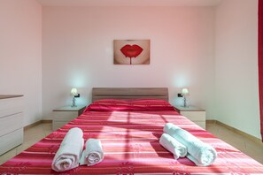 Habitación de hotel con cama, dos mesitas de noche con lámparas, un cuadro en la pared y dos toallas enrolladas sobre la cama.