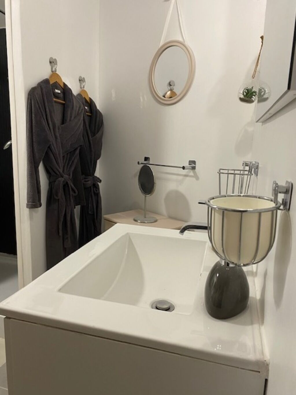 Un baño con un lavamanos blanco, un espejo y dos batas colgadas en un perchero.