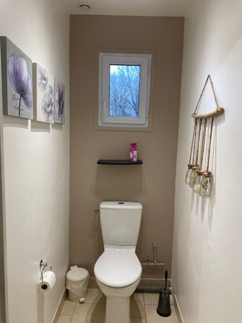 A small bathroom with a toilet, a bidet, a toilet paper holder, a window, and wall-mounted shelves.