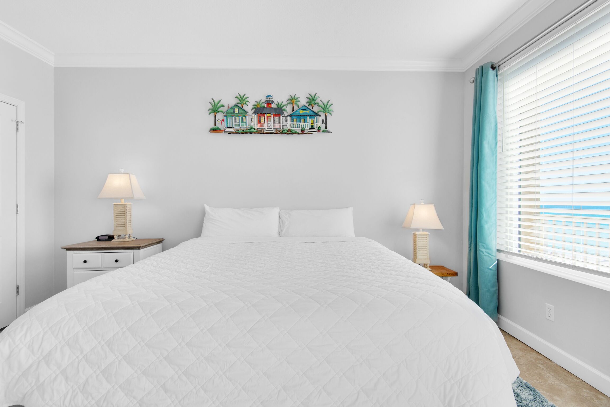 Une chambre avec un grand lit, deux tables de chevet avec des lampes, une œuvre d’art représentant une maison accrochée au mur, et une fenêtre avec des rideaux.