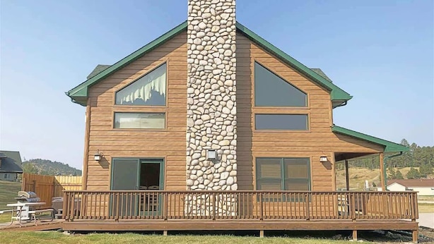 Una casa de madera de dos pisos con chimenea de piedra, ventanas grandes y una terraza de madera.