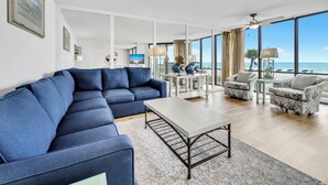 Uma sala de estar moderna com um sofá seccional azul, uma mesa de centro e vista para o oceano através de grandes janelas.