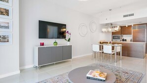 A modern living room with a flat-screen TV, a dining area with a table and chairs, and a kitchen with a microwave and refrigerator.