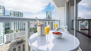 A balcony with a table set for breakfast, including a bowl of fruit, a glass of orange juice, and a champagne bottle.