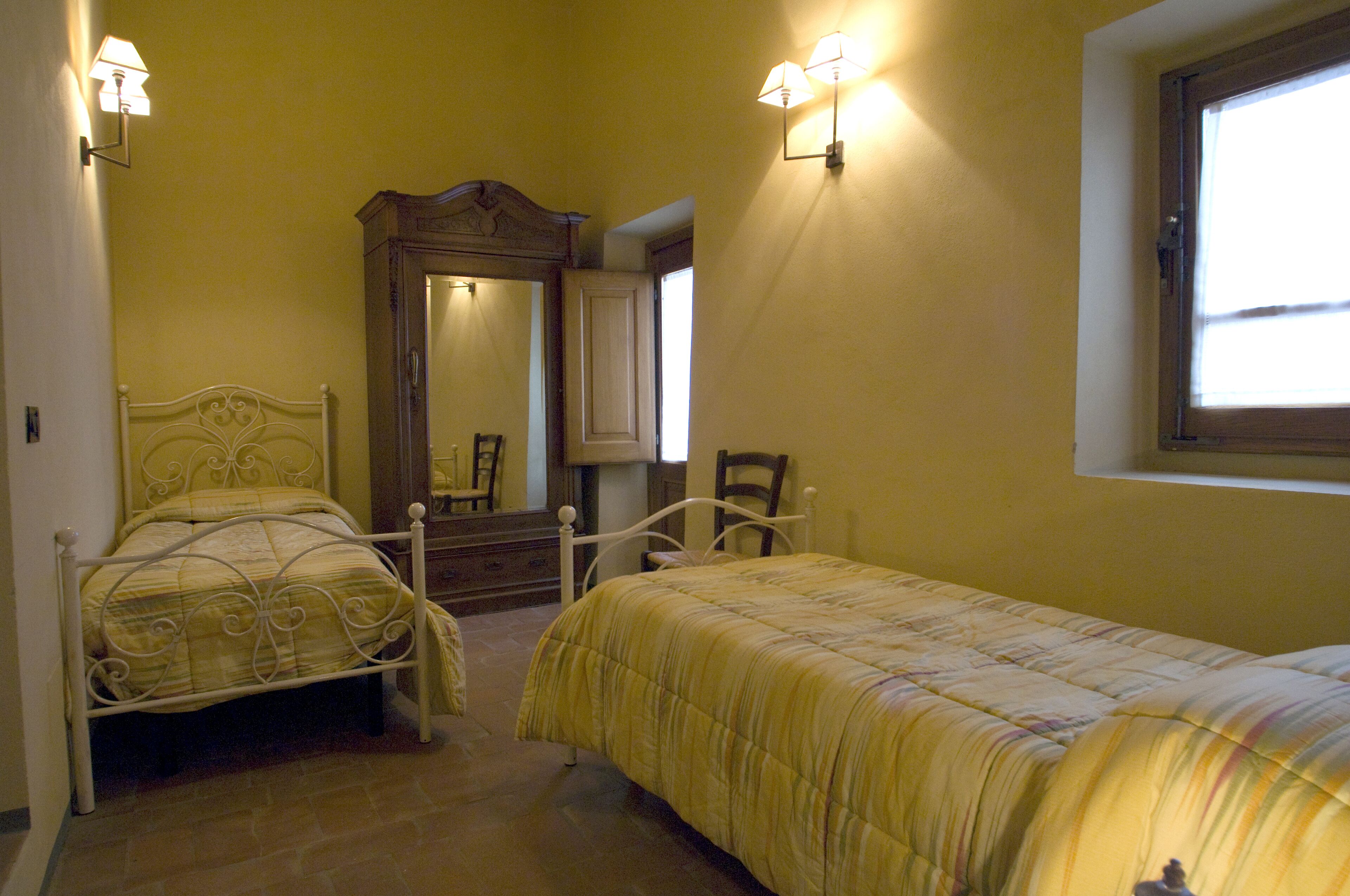 A bedroom with two beds, a wooden wardrobe, a mirror, and two wall-mounted lights.