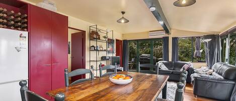 A dining area with a wooden table, chairs, and a bowl of fruit. There is a red door, a bookshelf, and a black leather sofa.