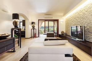 A modern living room with a stone wall, a flat-screen TV, a sofa, and a dining area with a wooden table. - Beautiful Luxury Villa Located in Sardinia in Villasimius Near the Beaches (Villasimius)