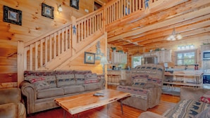 A wooden cabin interior with a staircase, two sofas with patterned cushions, a wooden coffee table, and a kitchen area with a dining table and chairs. - Majestic Splendor Lodge: Spacious deck, Home Theater, Game Room, Wooded View, near Attractions! (Sevierville)