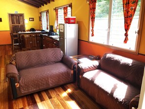 A cozy living room with a brown sofa, wooden flooring, and a kitchen area in the background. - 2BR Private Treetop Cottage With Bedroom AC in Tropical Gardens Near Town (San Ignacio)