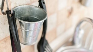 Ein Metall-Eimer hängt an einer Küchenwand, ein schwarzer Pfannenwender und ein Waschbecken mit Wasserhahn.