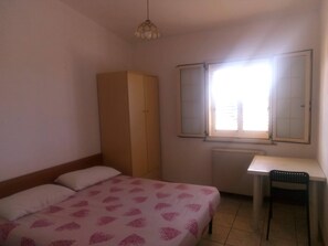 Ein Schlafzimmer mit Bett, Kleiderschrank, Schreibtisch und einem Fenster mit Jalousien.