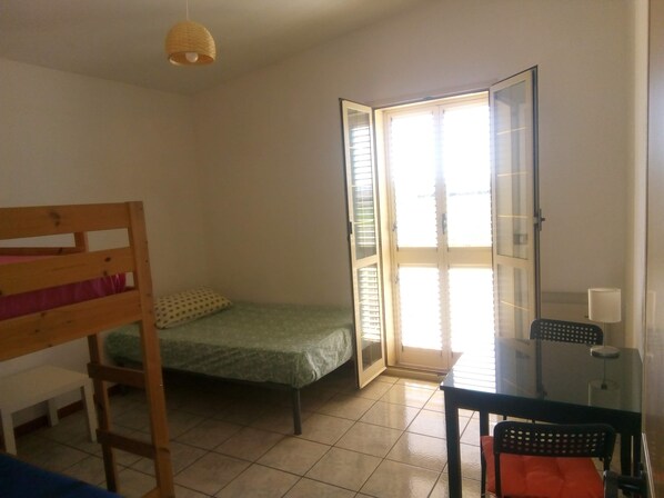Ein kleiner Raum mit einem Etagenbett, einem Einzelbett, einem Glastisch mit Stuhl und einer Tür zum Balkon.