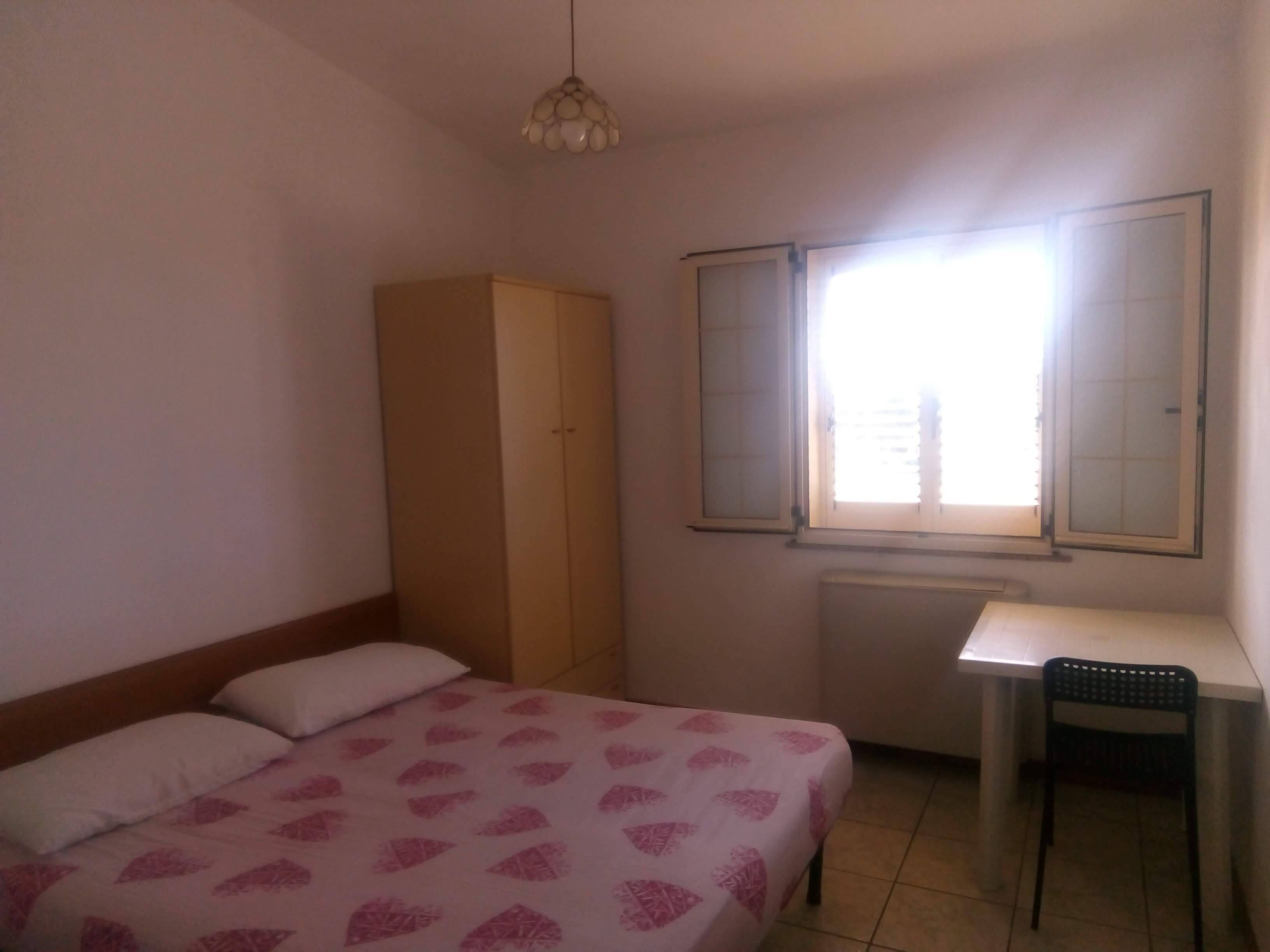 Ein Schlafzimmer mit Bett, Kleiderschrank, Schreibtisch und einem Fenster mit Jalousien.
