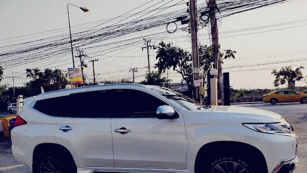 A white SUV parked on a street with overhead power lines and a yellow car in the background. - Room-thailand Taxi&apartment Hostel (Pak Kret)