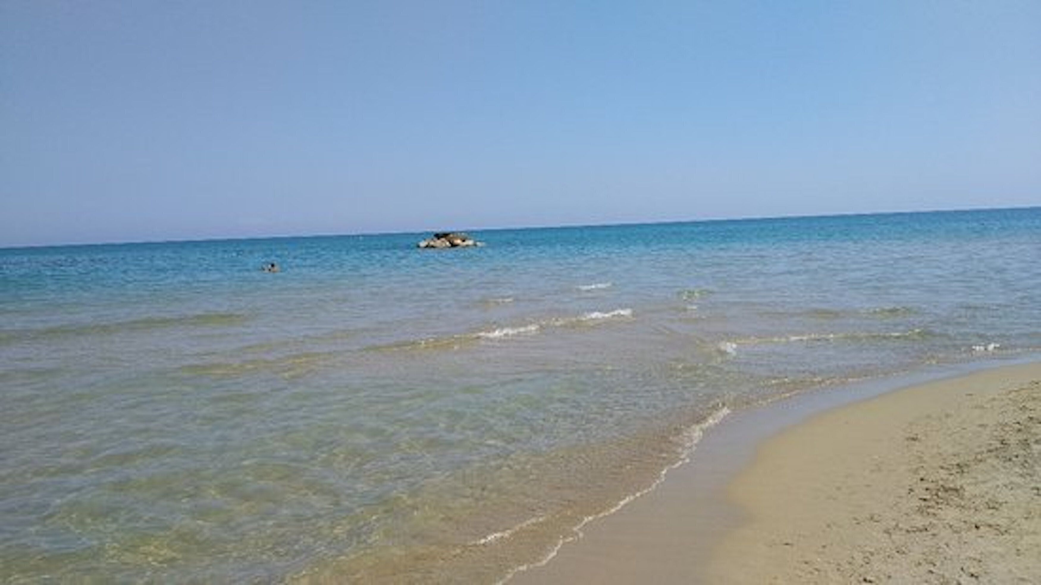 A beach with clear shallow waters, a sandy shore, and a distant rock formation.