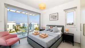 A bedroom with a large bed, a pink chair, a nightstand with a lamp, and a view of the city through a sliding door.