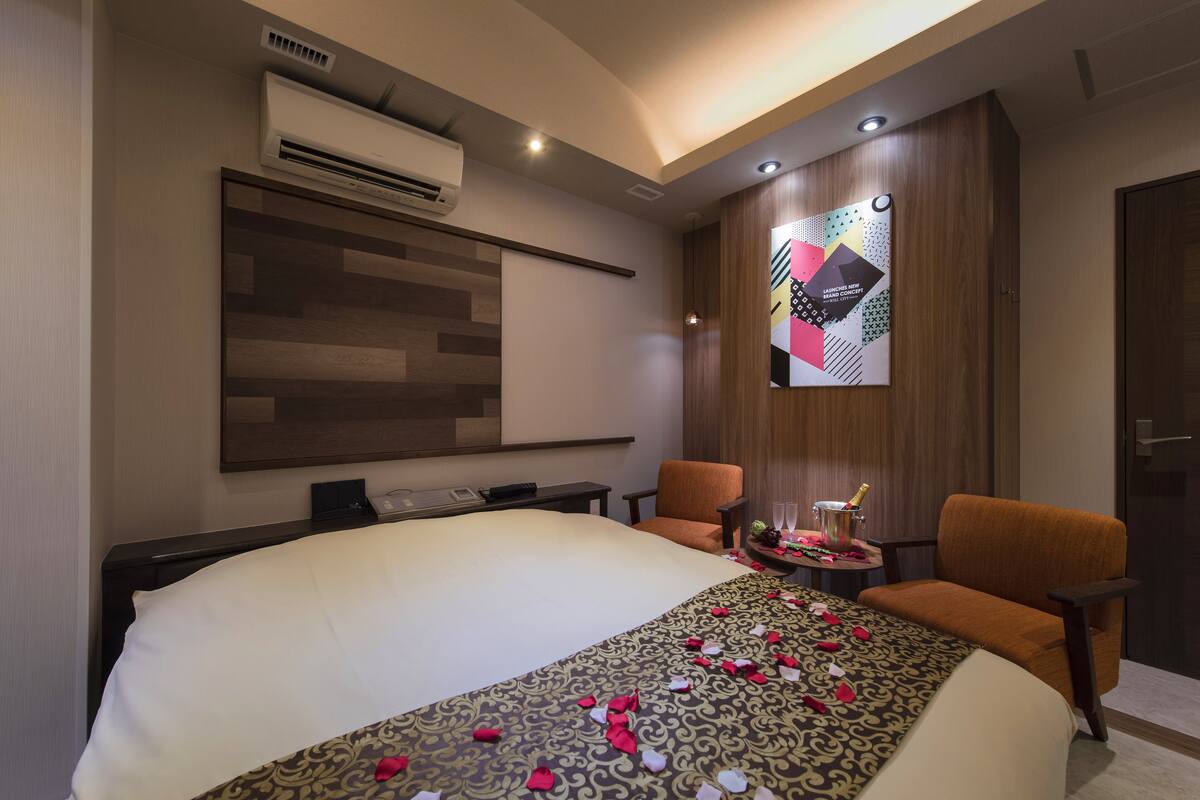 Una habitación de hotel moderna con una cama grande, dos sillones, una mesita con una bebida y un aire acondicionado de pared.