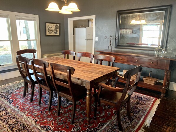 A dining area with a wooden table and chairs, a patterned rug, and a view of the outdoors through large windows.