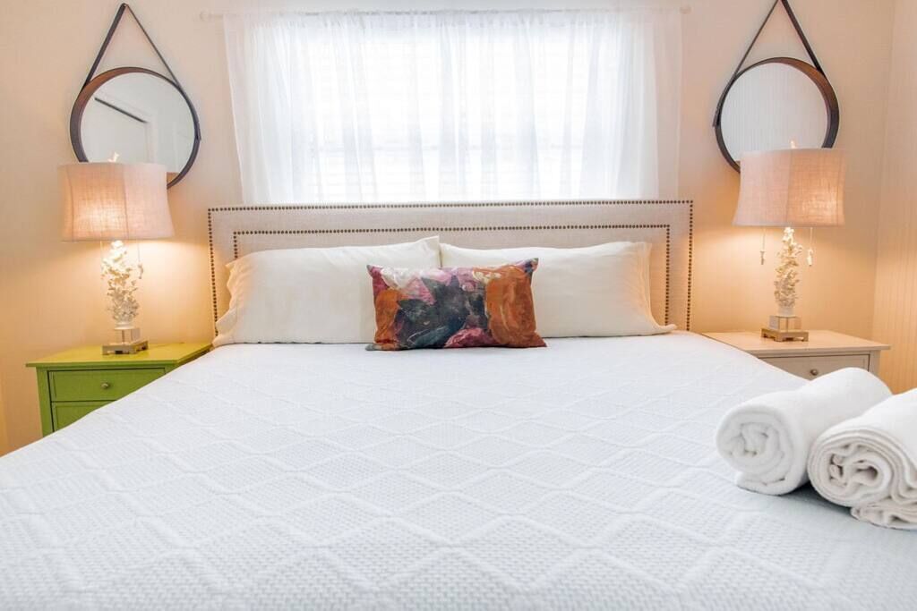 Un lit bien fait, recouvert d’une couette blanche, agrémenté d’un coussin coloré et de serviettes pliées, posés sur une table de chevet verte.