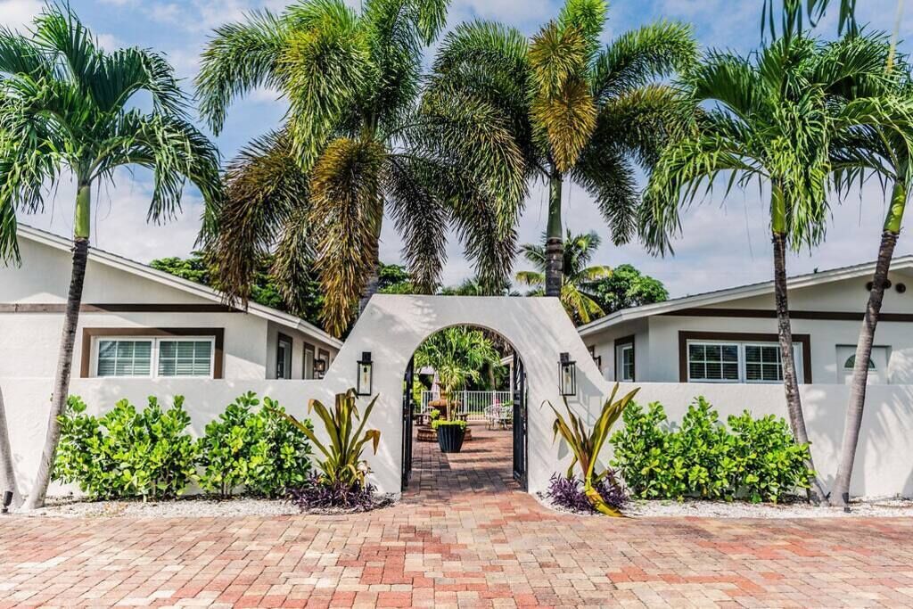 A white building with a brick pathway leading to an archway, surrounded by palm trees and greenery.