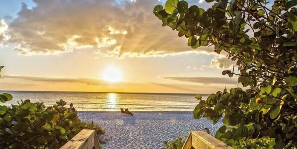 Ein Holzsteg führt zu einem Strand, im Hintergrund ein Sonnenuntergang.