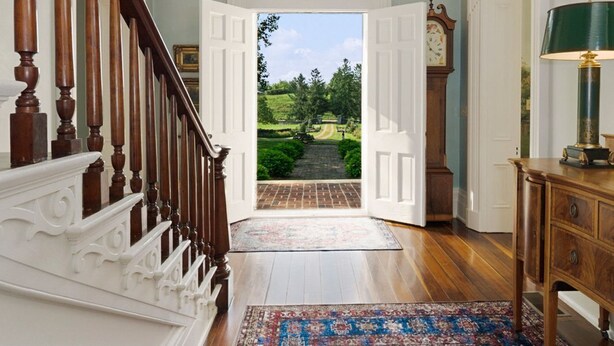 Un vaste hall d’entrée avec un escalier en bois, une grande moquette à motifs et une vue sur un jardin par une porte ouverte.