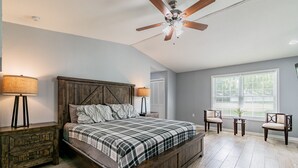 Une chambre avec un lit en bois, deux tables de chevet, un ventilateur de plafond, une fenêtre avec des stores et une petite table avec des chaises.