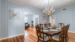 Une salle à manger avec une table en bois dressée pour six personnes, entourée de six chaises. Les murs de la pièce sont d’un bleu clair, le sol est en bois et un lustre illumine l’ensemble.