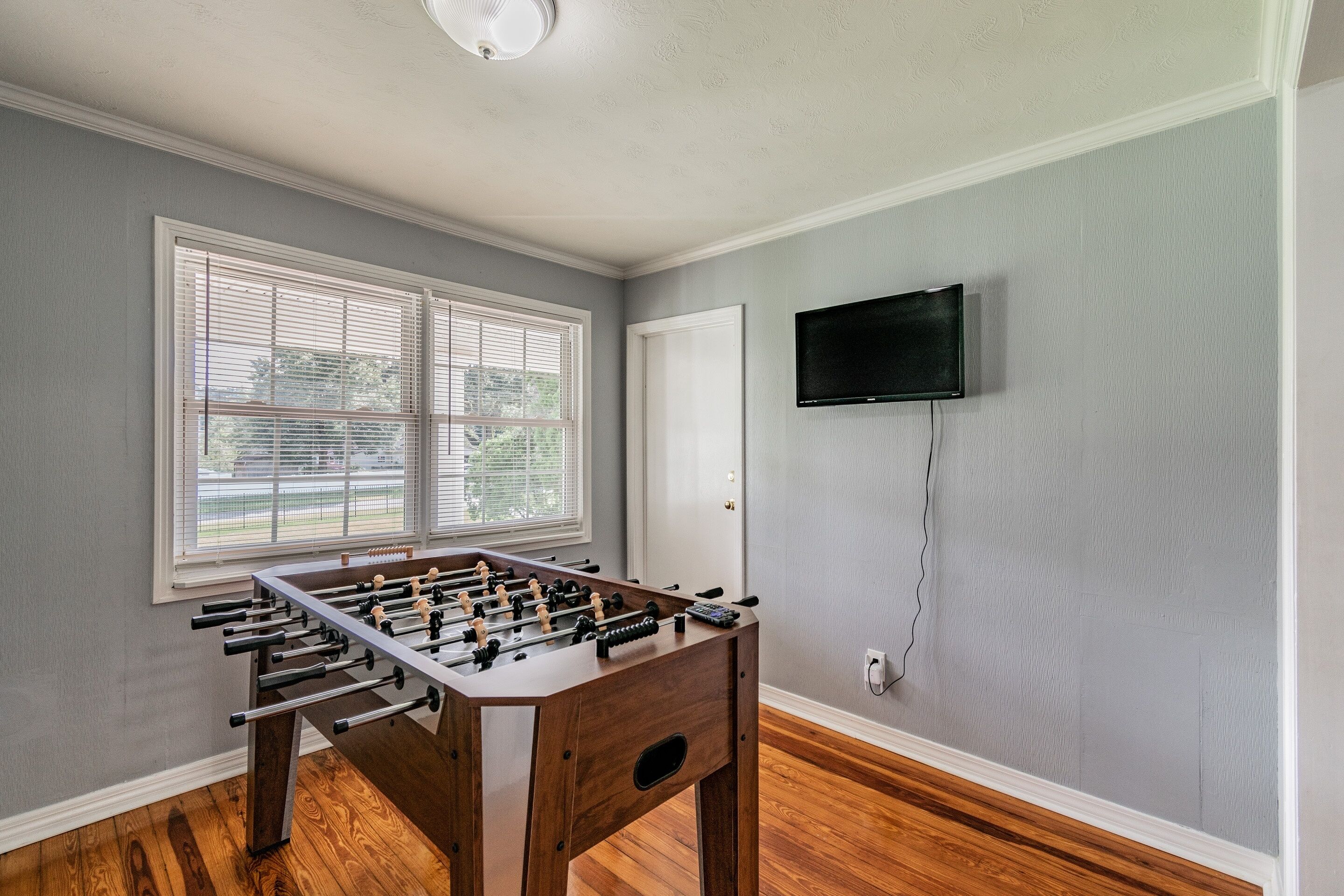 Una habitación con mesa de futbolín, televisión montada en la pared y ventana con persianas.