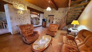 Ein gemütliches Wohnzimmer mit einer Steinwand, Holzbalkendecke und einer Treppe. Zwei Ledersessel, ein Holztisch und eine Lampe auf einem Beistelltisch sind vorhanden.