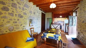 A cozy room with a stone wall, wooden ceiling beams, a yellow sofa, a table with a quilt, and a kitchen area visible through an open door. - Gîte Nico Label Pêche - 4 pers proche du Gers (LANNES)
