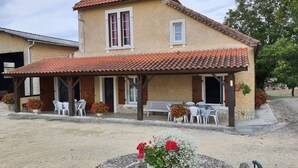 A two-story house with a tiled roof, a covered patio with outdoor seating, and a small garden area with flowers. - Gîte Nico Label Pêche - 4 pers proche du Gers (LANNES)