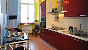 A kitchen with red cabinets, a white countertop, and a tiled floor. There is a window with curtains, a chair, and a table with a vase and a teapot. - Uno sguardo su Porta Palazzo (Turin)