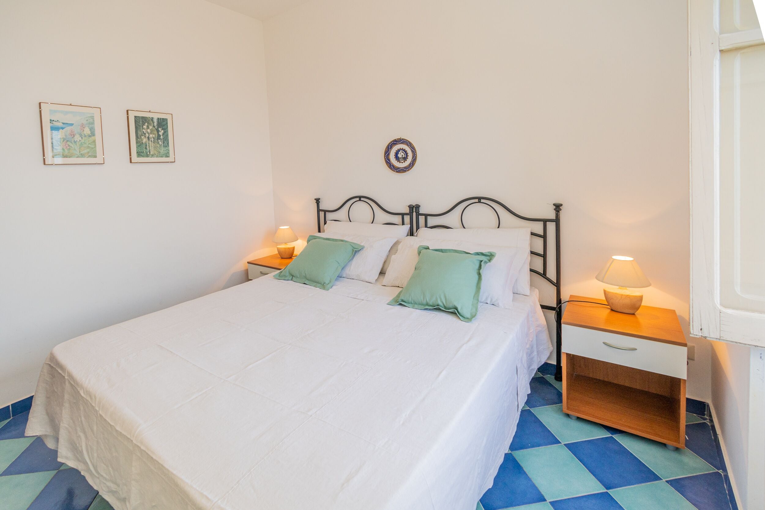 A neatly made bed with a white bedspread and two pillows, a bedside table with a lamp, and a wall with two framed pictures.