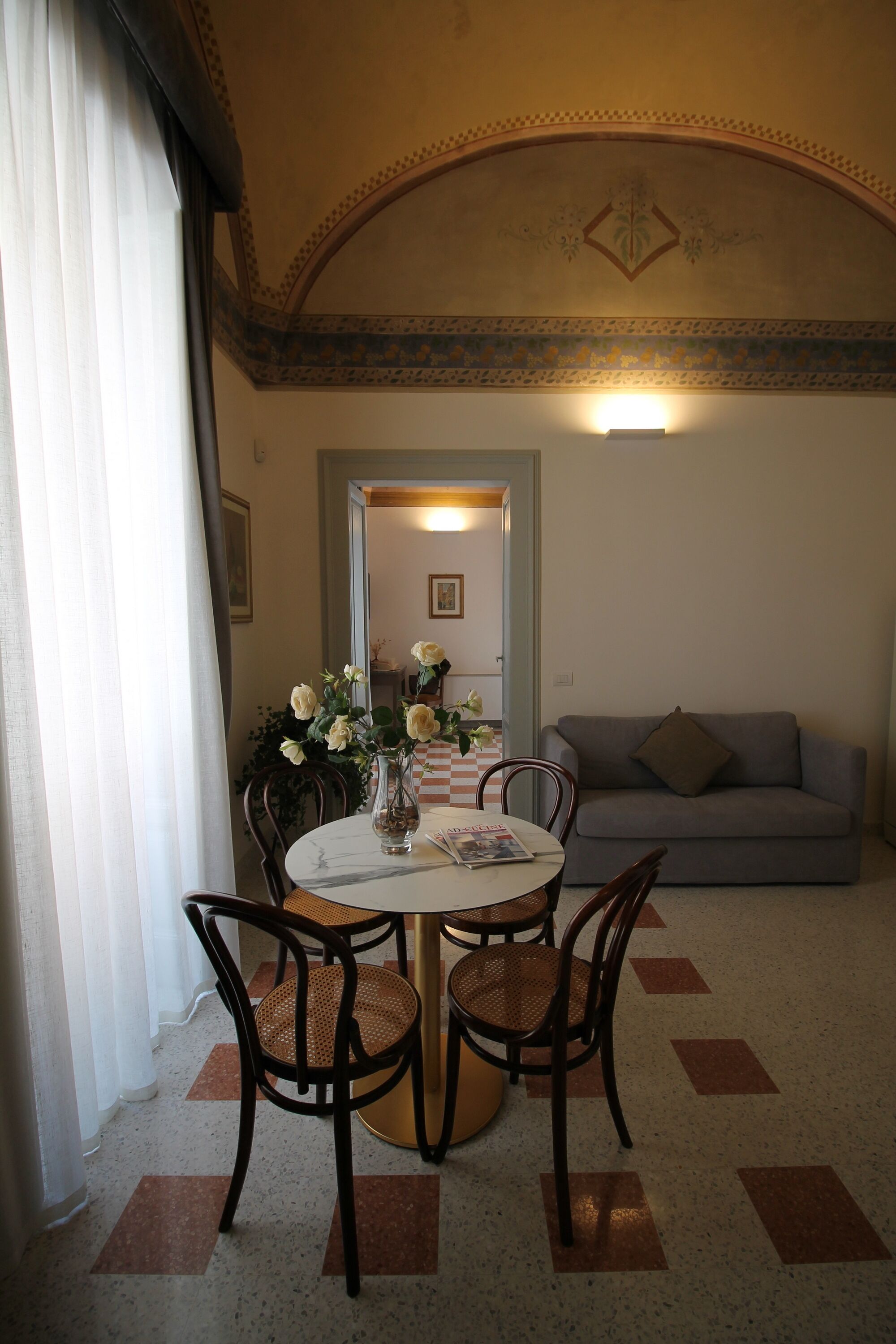 A dining area with a round table, four chairs, a vase with flowers, and a grey sofa in the background.