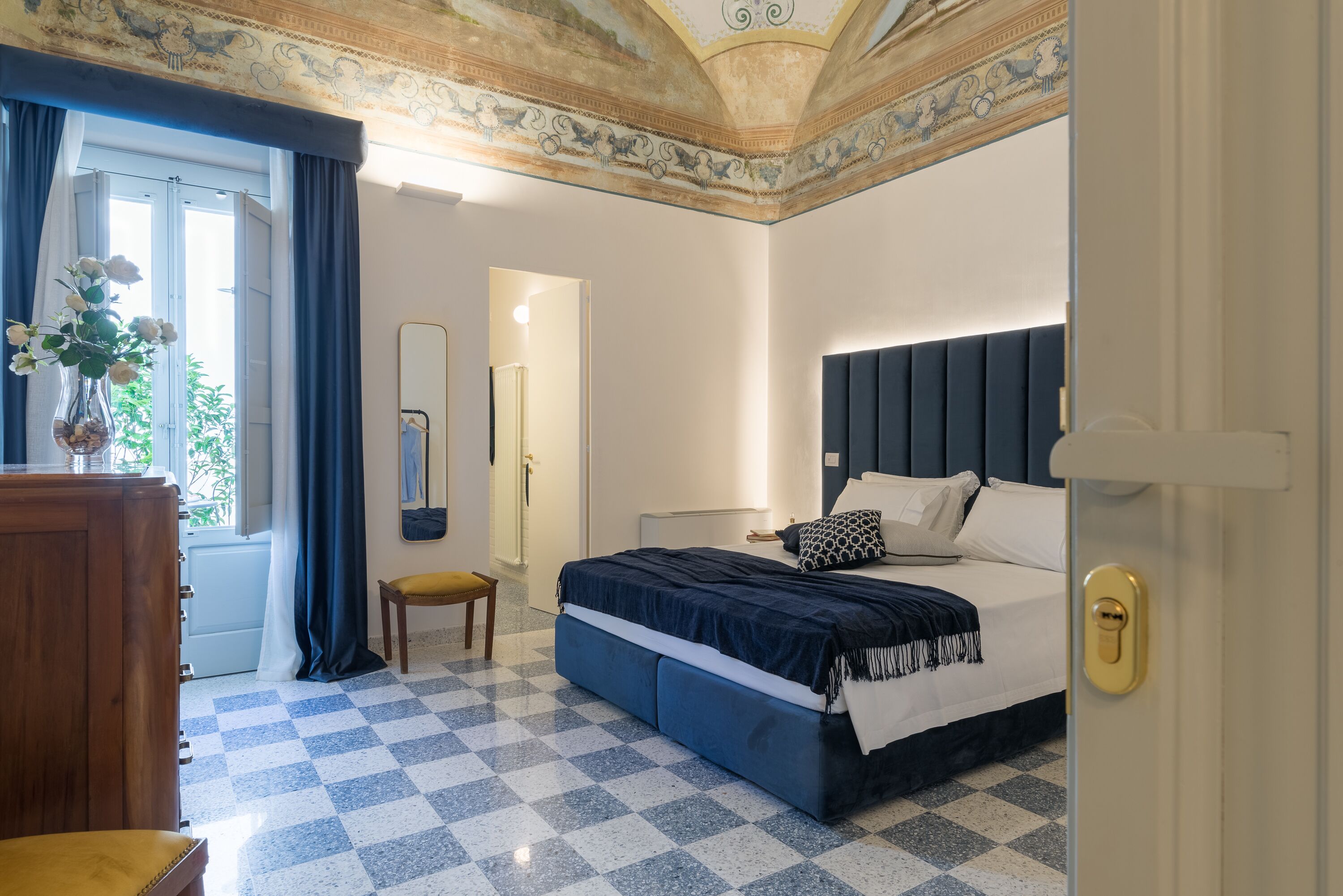 Une chambre d’hôtel avec un sol à carreaux, un lit doté d’une tête de lit sombre, une commode en bois et une fenêtre avec des rideaux.