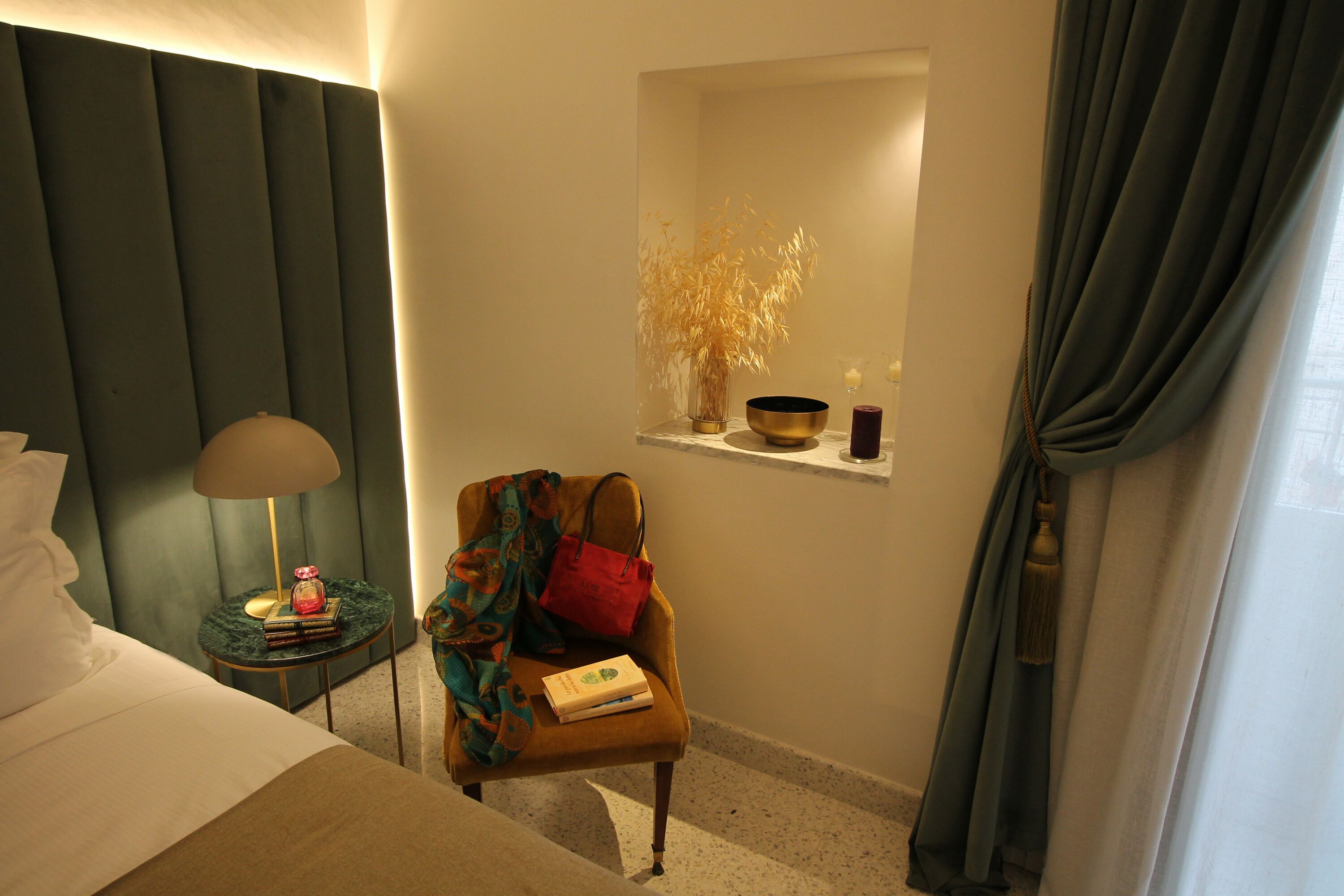 A hotel room with a bed, a nightstand with a lamp, a chair with a book, a small shelf with decorative items, and a window with curtains.