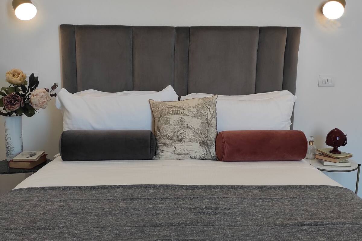 Un lit bien fait, avec une tête de lit grise, des oreillers blancs et un coussin décoratif représentant un paysage naturel.