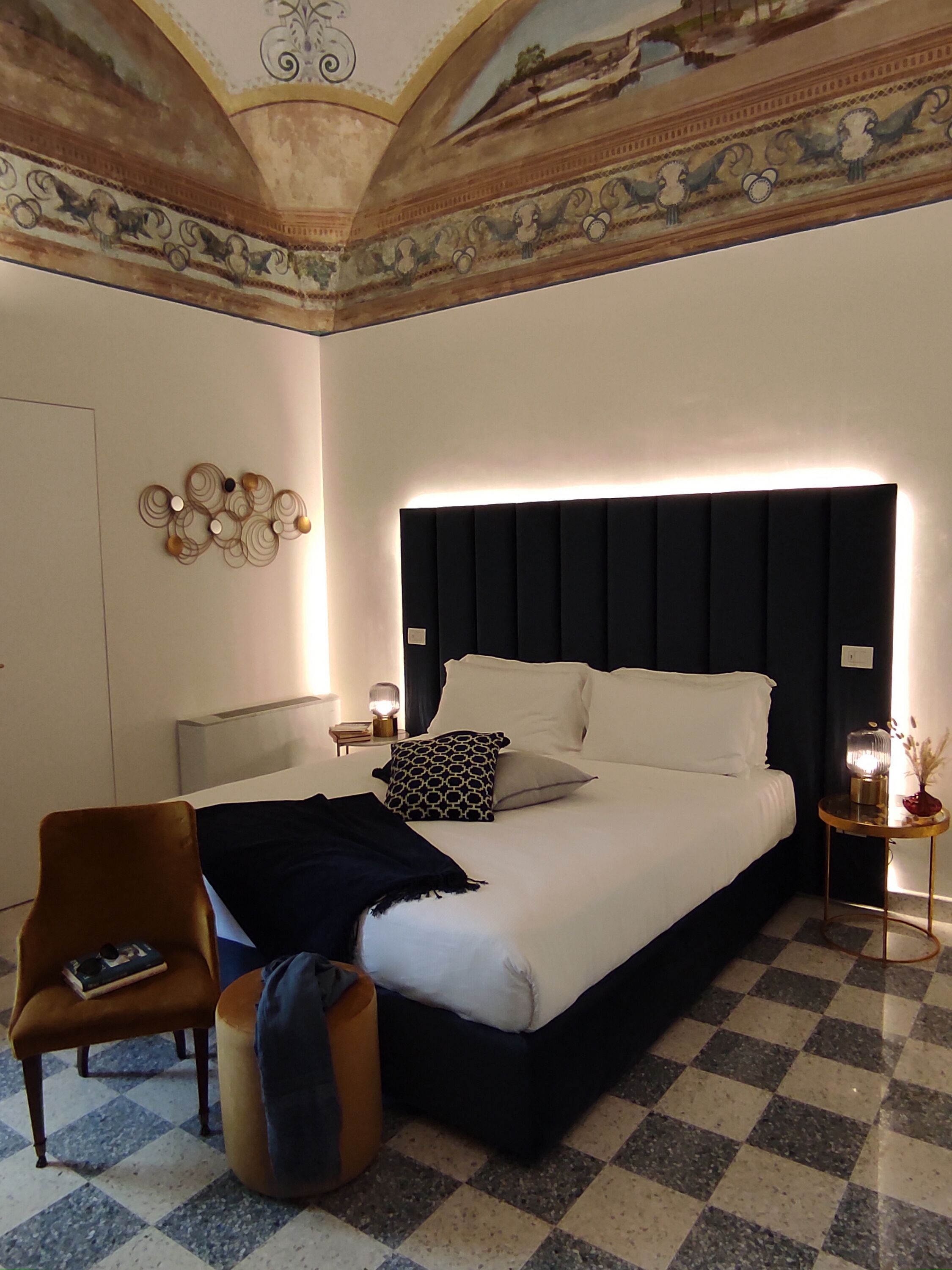 A hotel room with a large bed, a chair, a small table with a lamp, and a decorative wall piece.