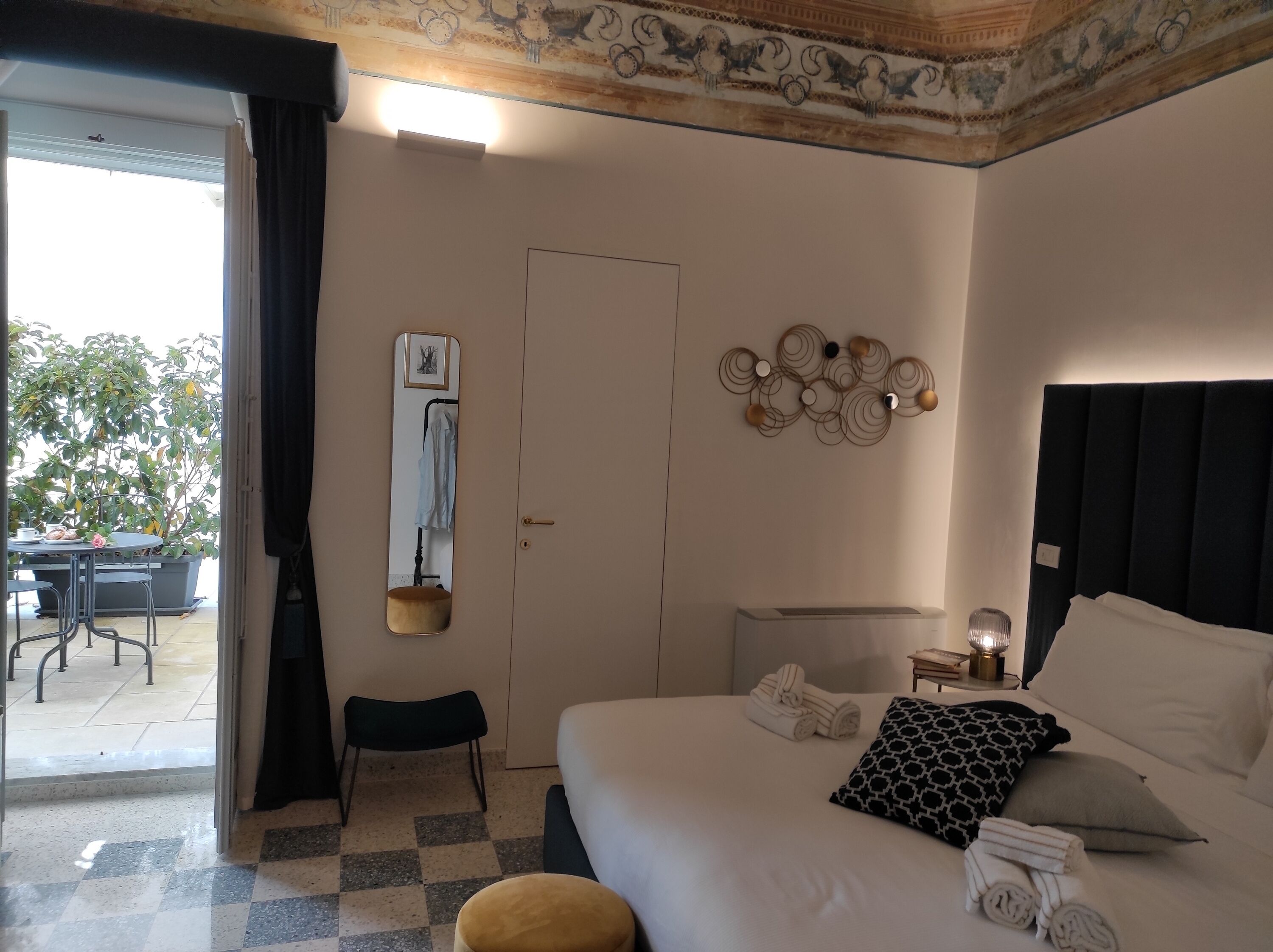 Une chambre d’hôtel comprenant un lit, une table de chevet avec une lampe, un miroir et une vue sur un espace extérieur aménagé avec une table et des chaises.