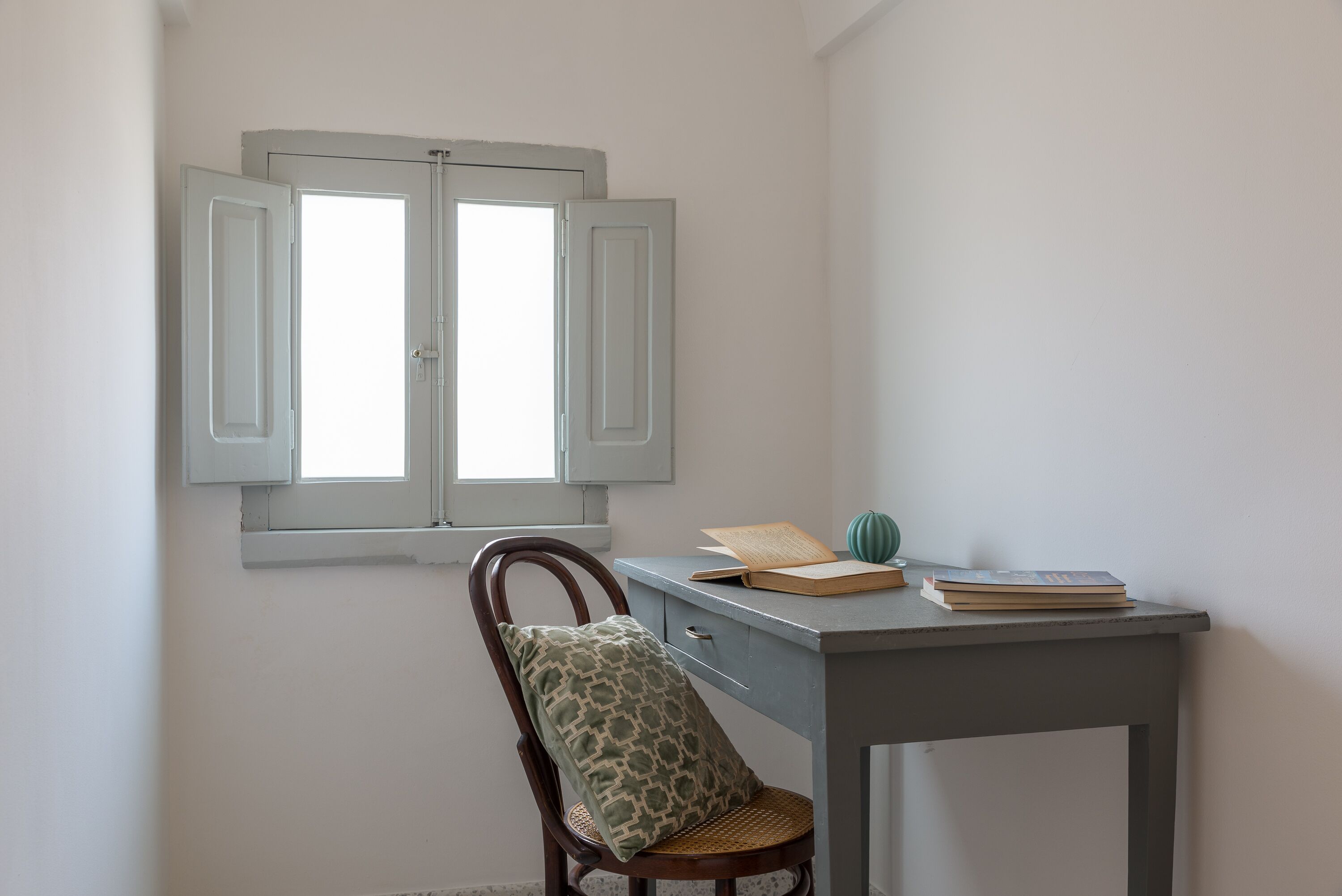 A room with a desk, a chair, a window with shutters, and a cushion on the chair.
