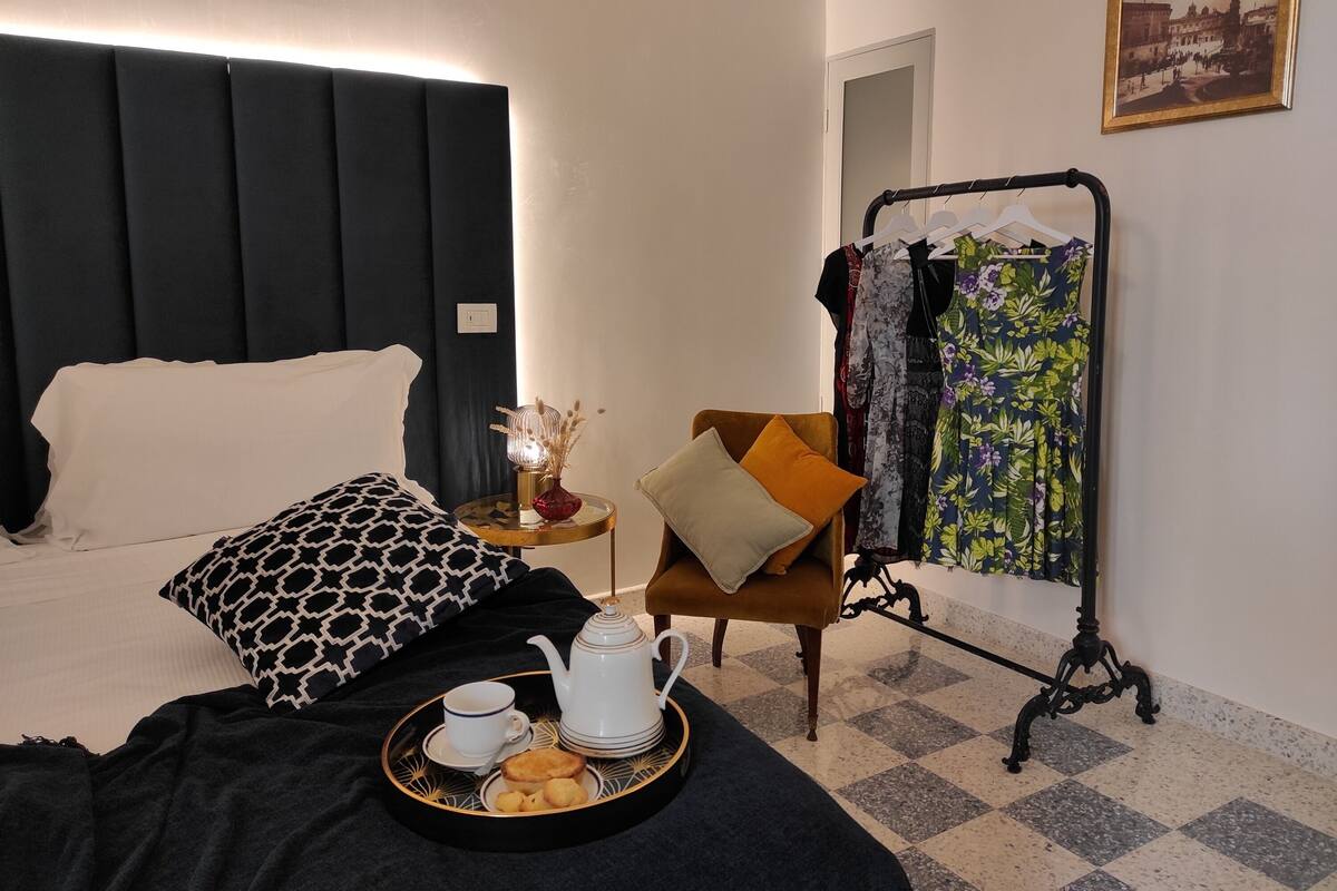 Une chambre d’hôtel avec un lit, une table de chevet sur laquelle se trouvent une théière et une assiette de biscuits, une chaise, un porte-vêtements et un tableau encadré au mur.