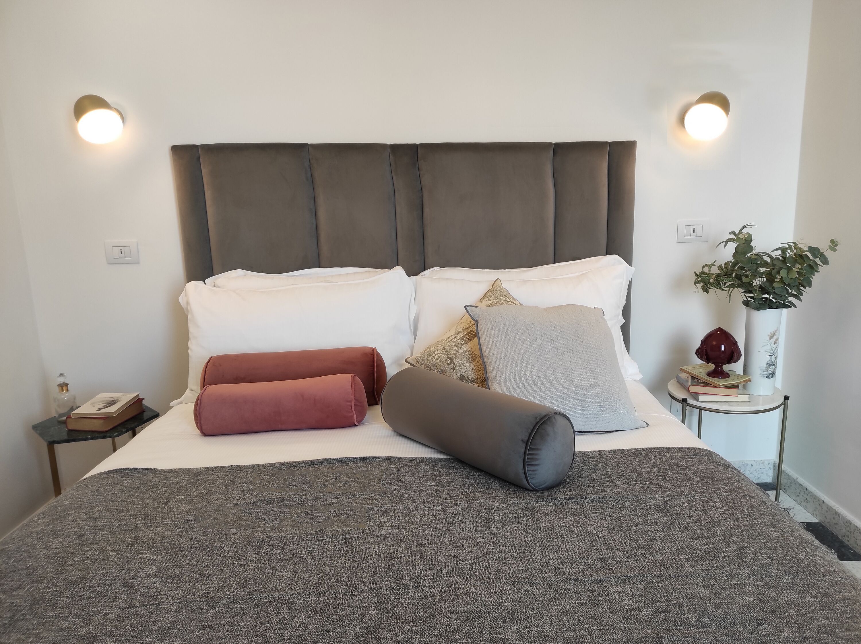 Una cama bien hecha, con cabecera gris, almohadas blancas y manta gris. Se ve una mesita de noche con un libro y una botella.