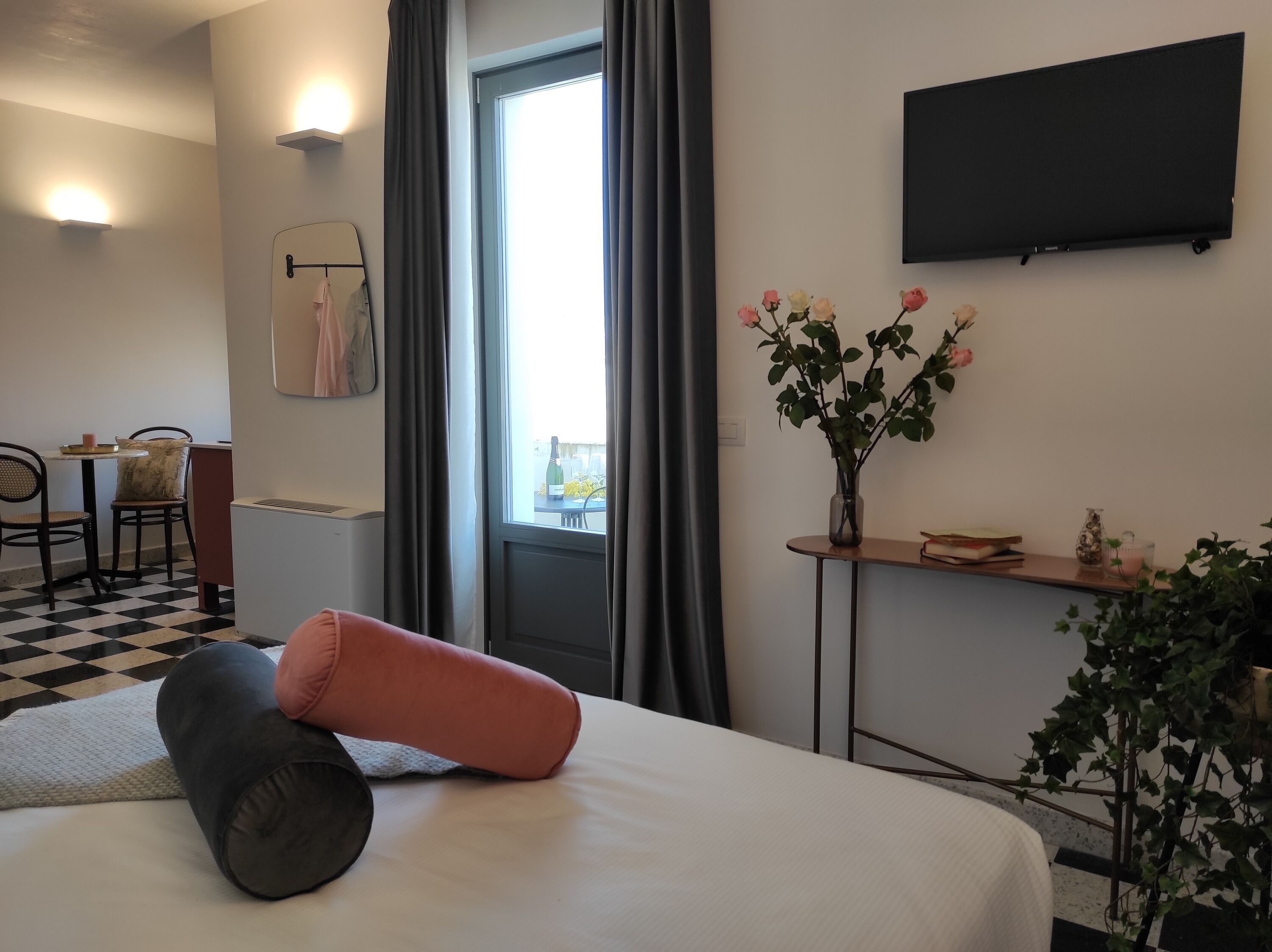 A hotel room with a bed, a desk with a vase of flowers, a flat-screen TV, and a window with curtains.