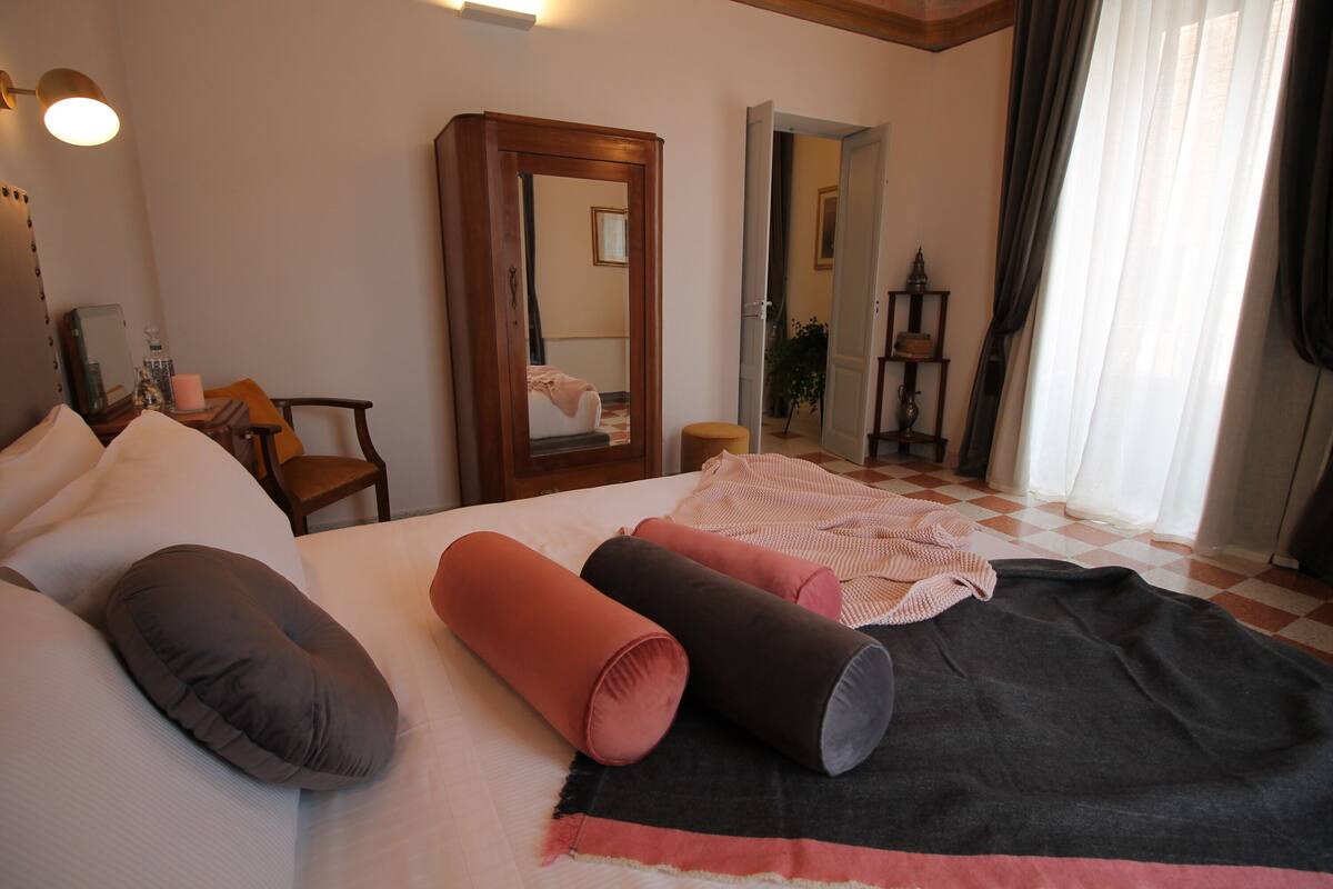 Une chambre d’hôtel avec un lit, des oreillers, une couverture, un miroir, une chaise et une petite table avec une lampe.