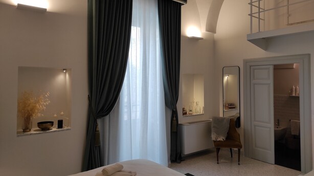 A modern hotel room with a large bed, a sitting area with a chair, a bathroom visible through an open door, and a balcony with a railing. - 1940 Luxury Accommodations by Wonderful Italy (Ostuni)