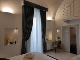 A modern hotel room with a large bed, a sitting area with a chair, a bathroom visible through an open door, and a balcony with a railing.